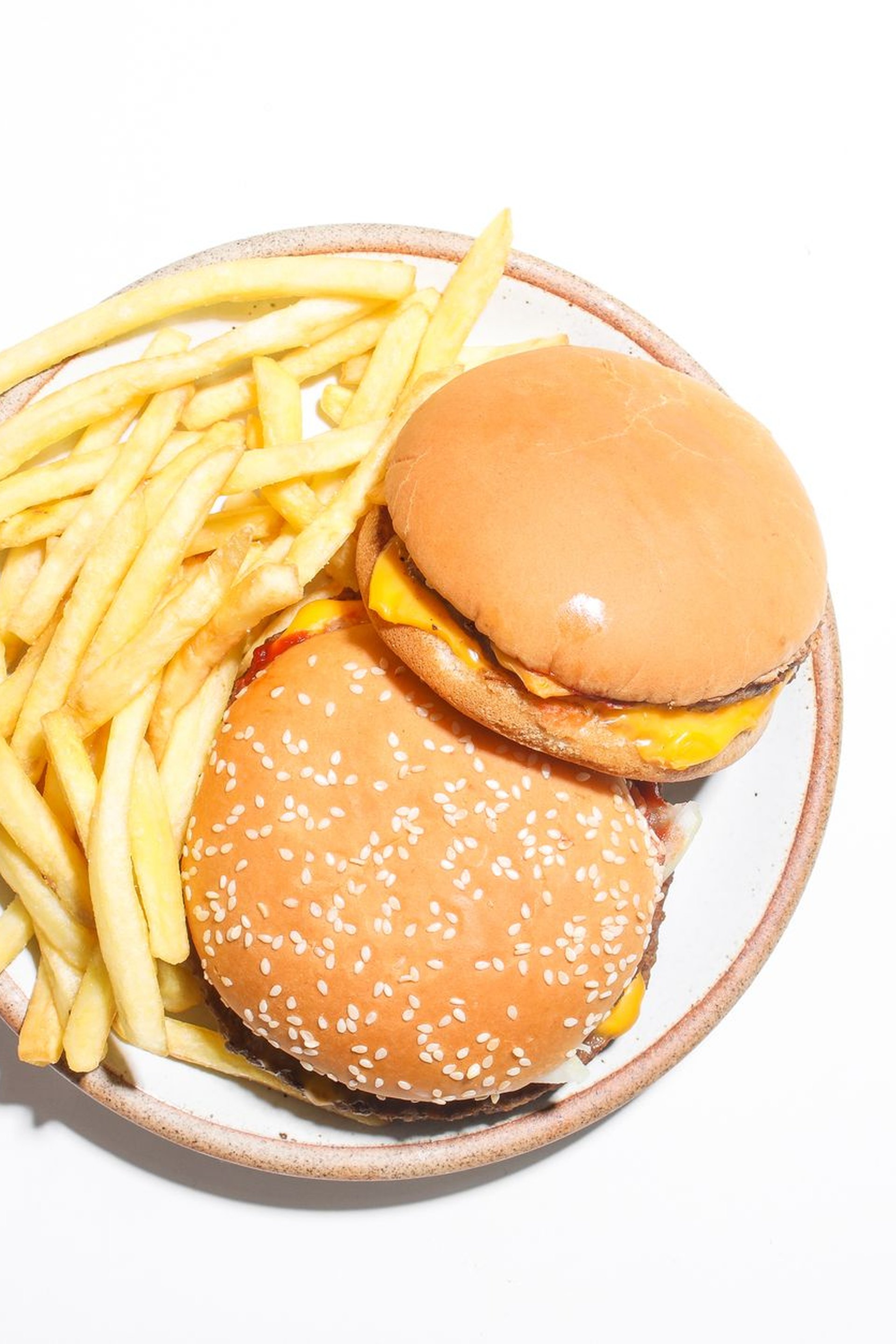 Vidám fiatal nő két hamburgert tart az arca mellé és mosolyog egy beltéri fal előtt.